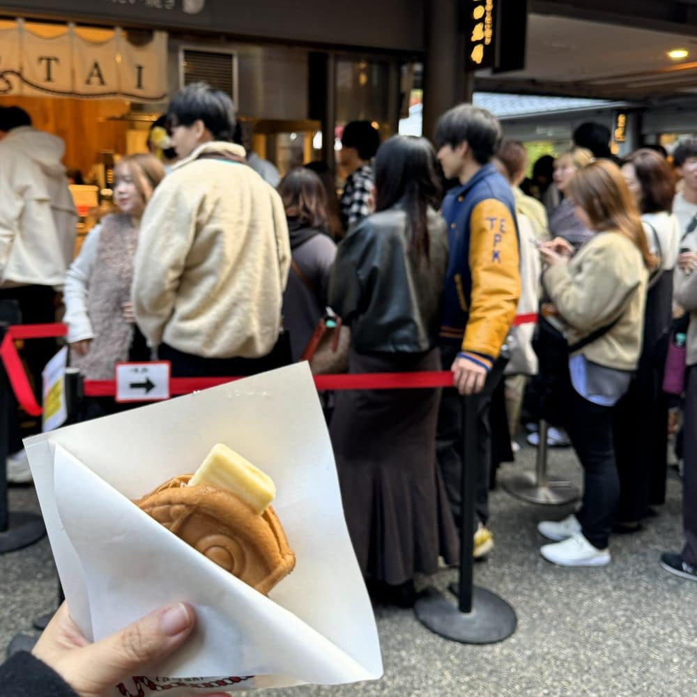 Freshly baked round red bean and butter taiyaki (An-Butter Taiyaki) held in front of a busy customer queue outside Mamemono to Taiyaki in Arashiyama, Kyoto, Japan.