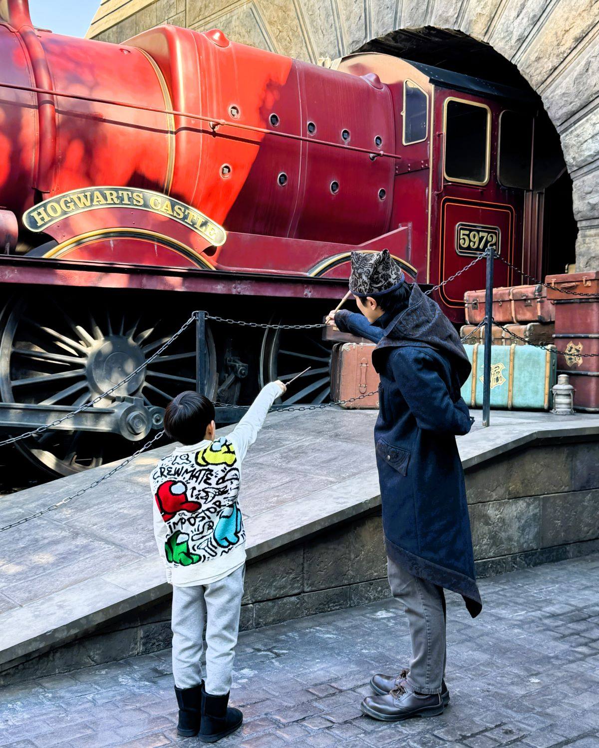 日本大阪環球影城(USJ)活米村車站旁,一名孩子與列車員並排,手持魔杖施法。