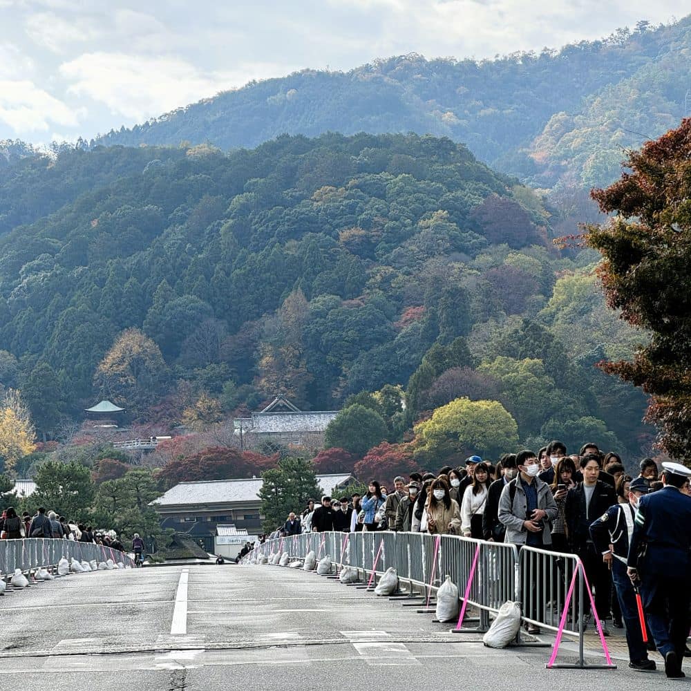 Crowd control barricades and tourists walking across Togetsukyo Bridge in Arashiyama, Kyoto, Japan, set against autumn foliage-covered mountains.