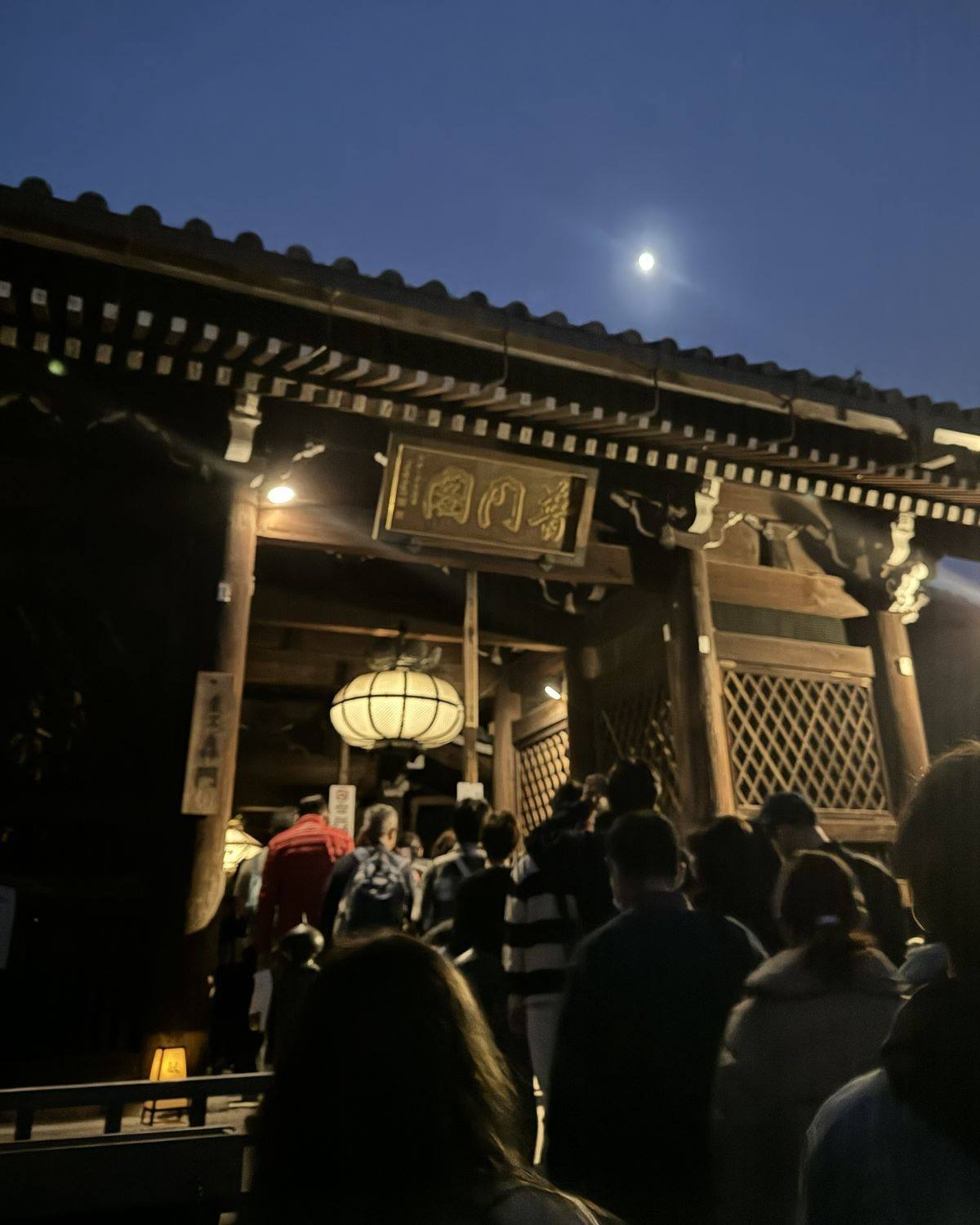 Visitors entering an illuminated traditional wooden gate with a paper lantern under a moonlit night sky at Kiyomizu-dera Temple in Kyoto, Japan.