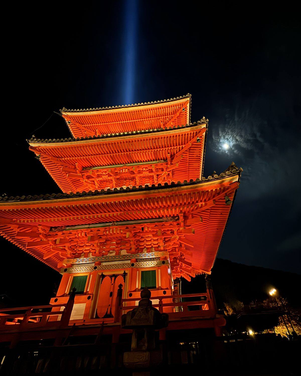 Illuminated three-story vermilion pagoda (Sanjunoto) with a vertical blue light beam and moon during special night viewing at Kiyomizu-dera Temple in Kyoto, Japan.