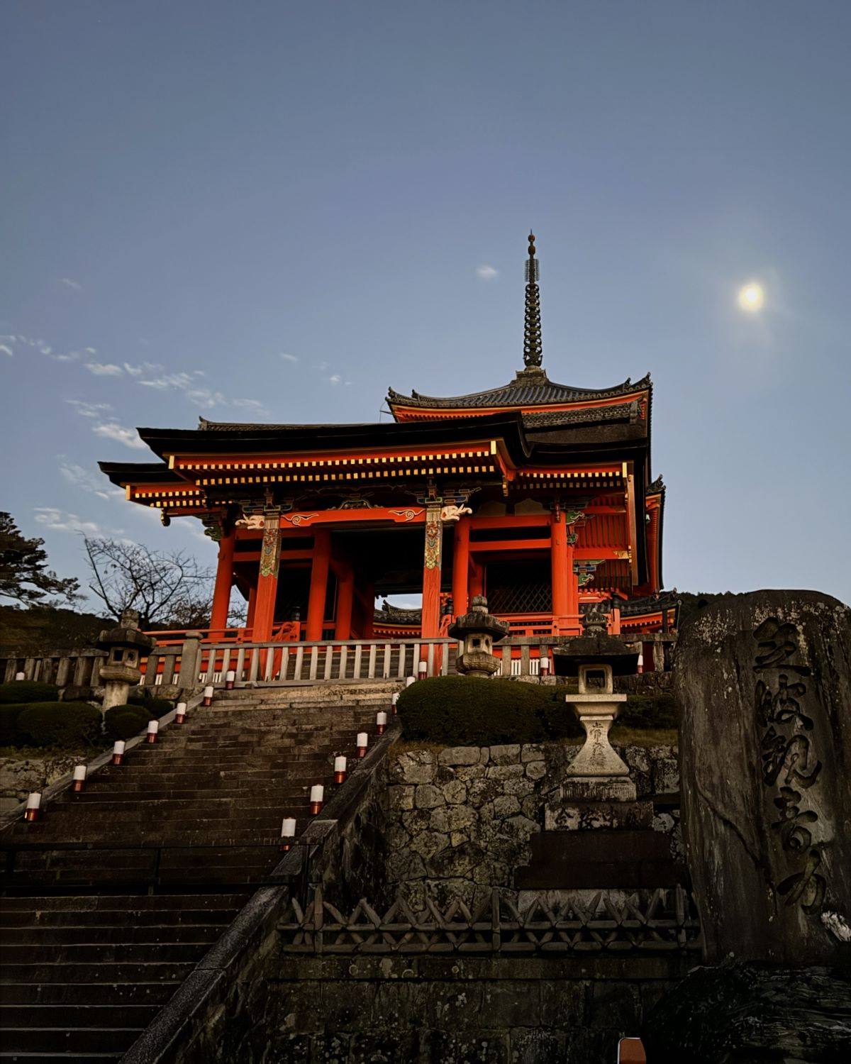 The red Niomon Gate and stone staircase lined with lanterns under a full moon at twilight at Kiyomizu-dera Temple in Kyoto, Japan.