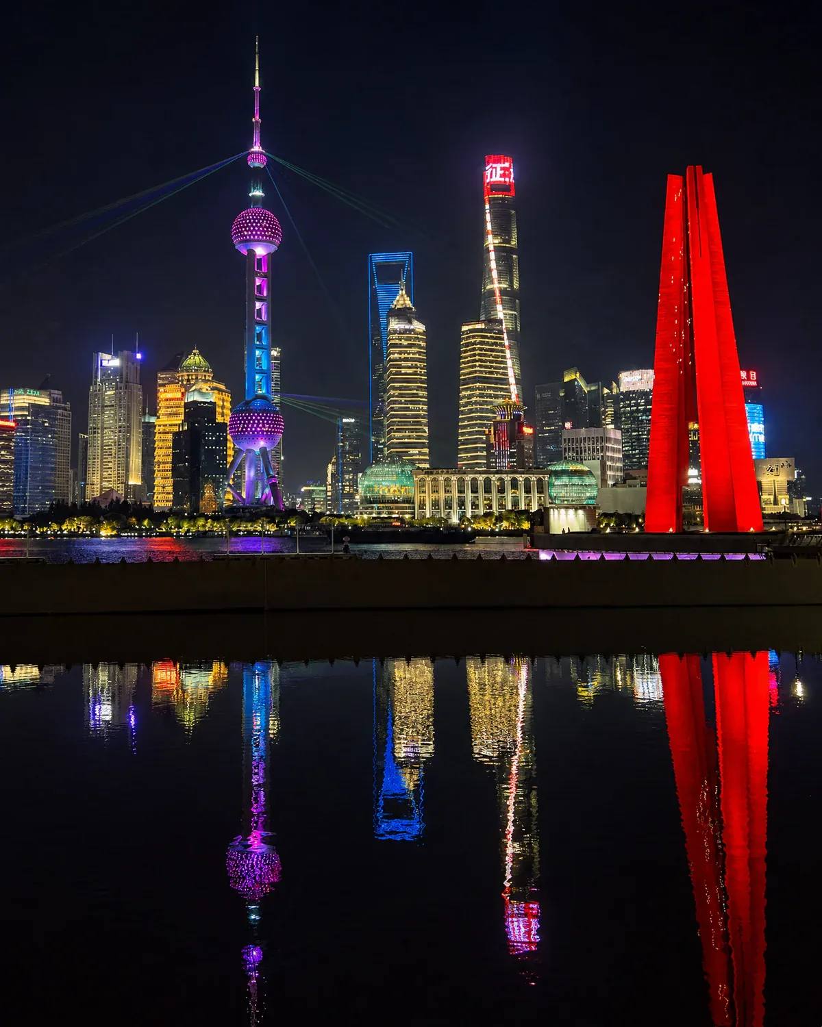 Illuminated Lujiazui skyline at night featuring Oriental Pearl Tower and Shanghai Tower reflected in the river in Shanghai, China.
