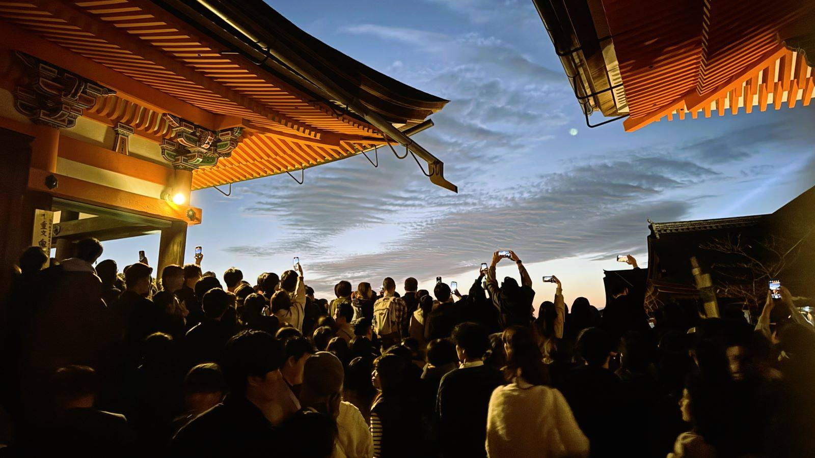 Crowd holding up smartphones to photograph the twilight sky beneath illuminated vermilion temple eaves at Kiyomizu-dera Temple in Kyoto, Japan.