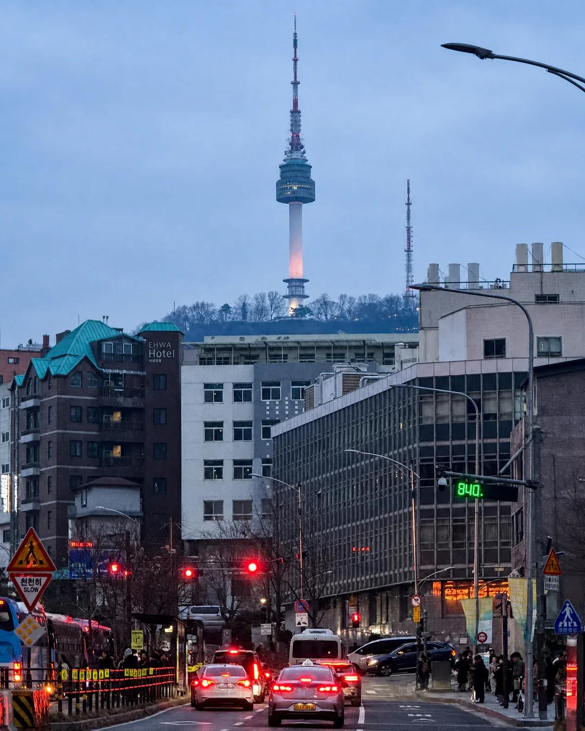 Street view of the illuminated N Seoul Tower on Namsan Mountain behind city buildings at dusk in Seoul, South Korea.