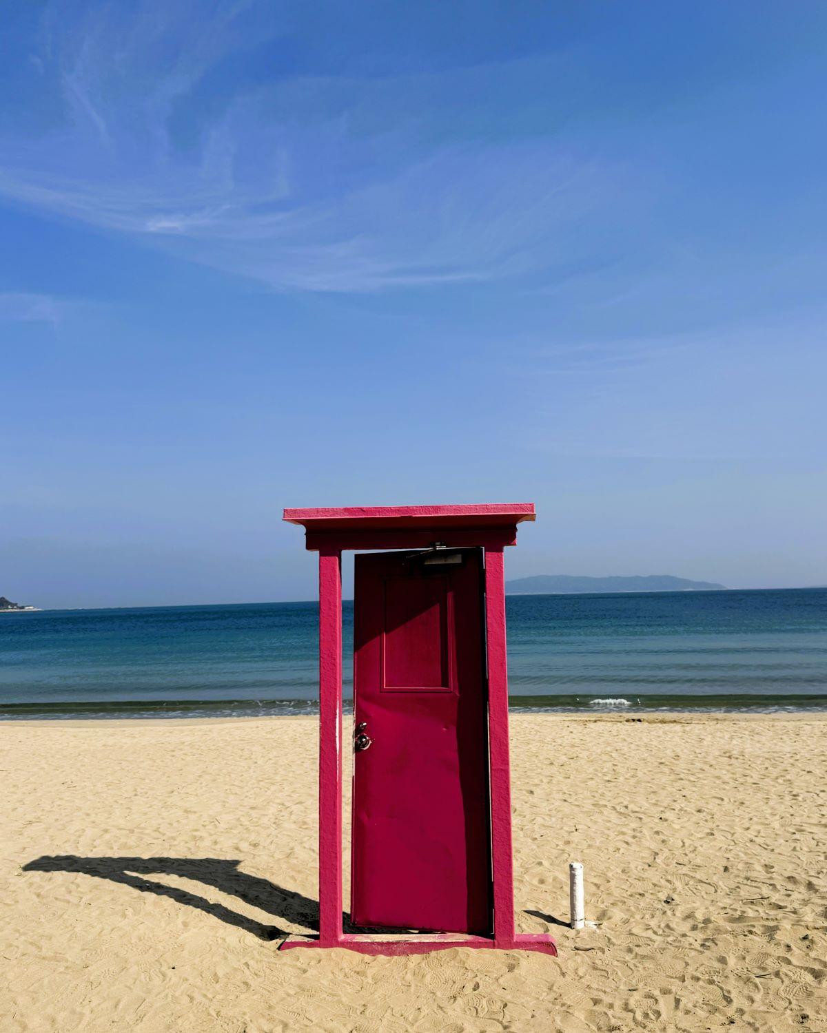 The vibrant pink "Anywhere Door" (Dokodemo Door) photo installation standing on the sandy beach along the Itoshima coastline in Fukuoka Prefecture, Japan.