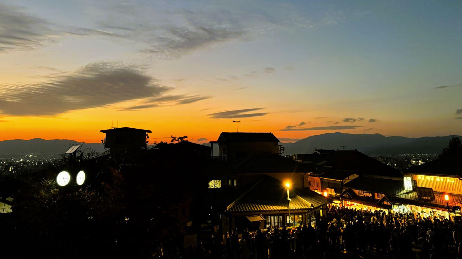 Vibrant orange sunset over traditional wooden shop roofs and crowds along the approach to Kiyomizu-dera Temple in Higashiyama, Kyoto, Japan.